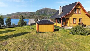 Une maison jaune avec un toit noir, un petit abri de jardin et un mât sur une pelouse, avec un lac et des collines en arrière-plan.
