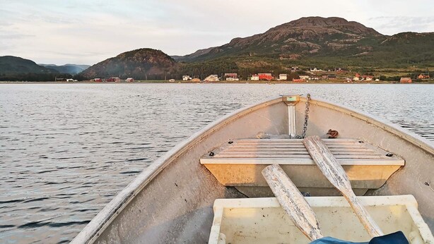 Ein Boot mit einer hölzernen Bank und einer Kette, auf ruhigem Wasser mit einer Stadt und Bergen im Hintergrund.