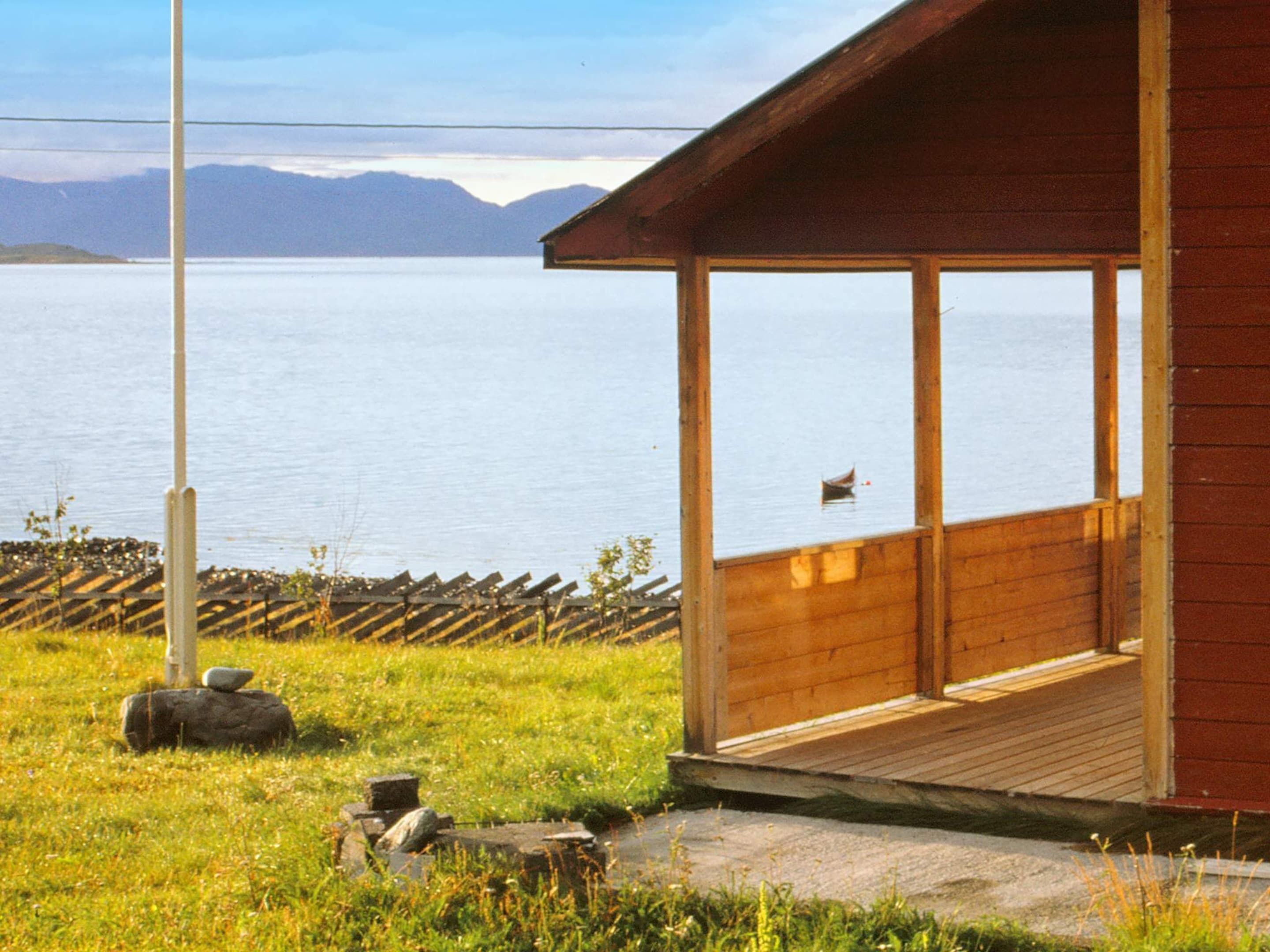 A wooden cabin with a porch overlooking a calm lake and mountains in the distance.