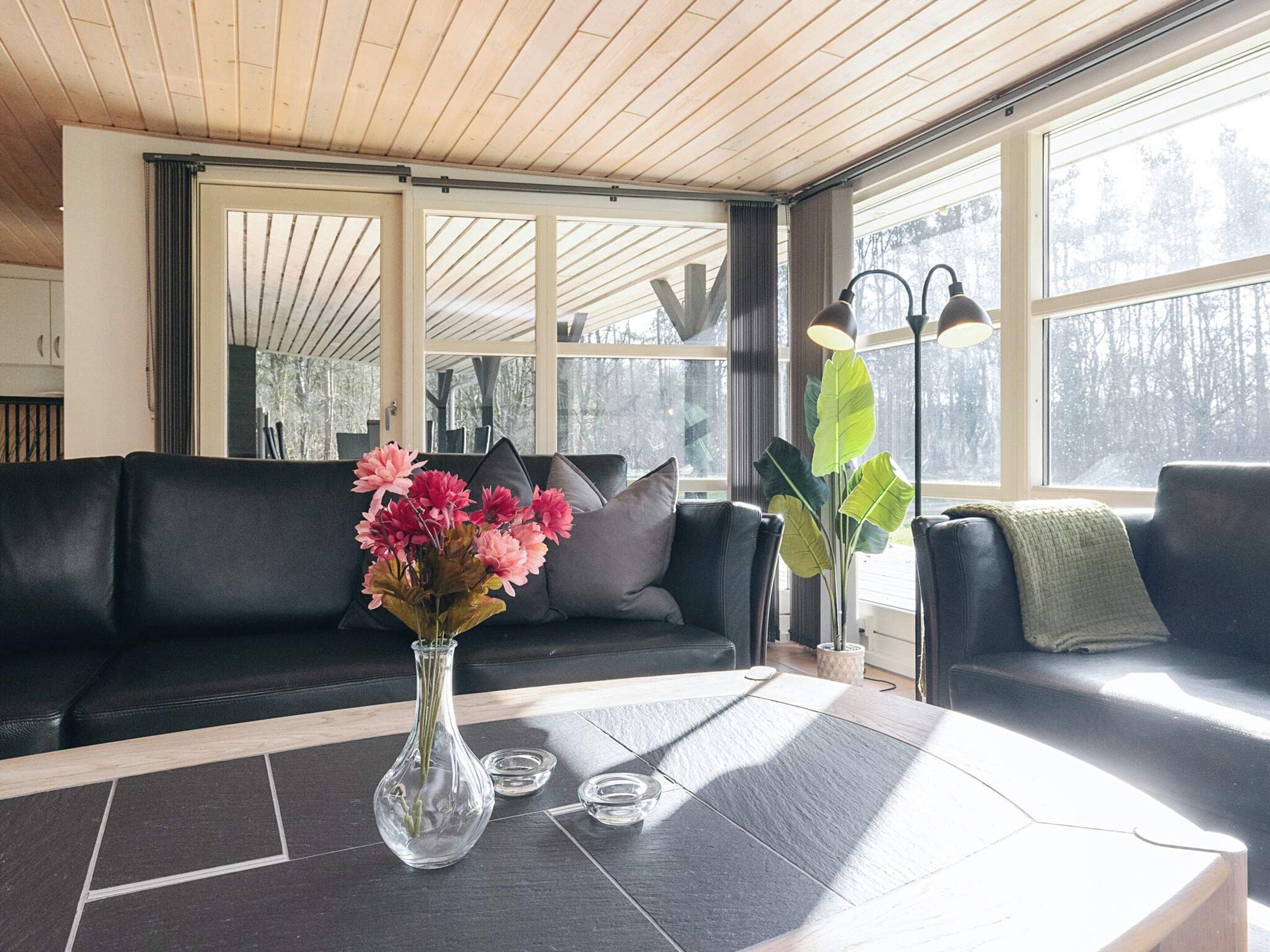 A modern living room with a black leather sofa, a glass vase with flowers, and a large window with a view of trees.