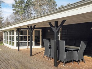 A covered outdoor dining area with a table and chairs, a wooden deck, and a building with large windows and a dark exterior. - 5 Star Holiday Home in Hals (Asaa)