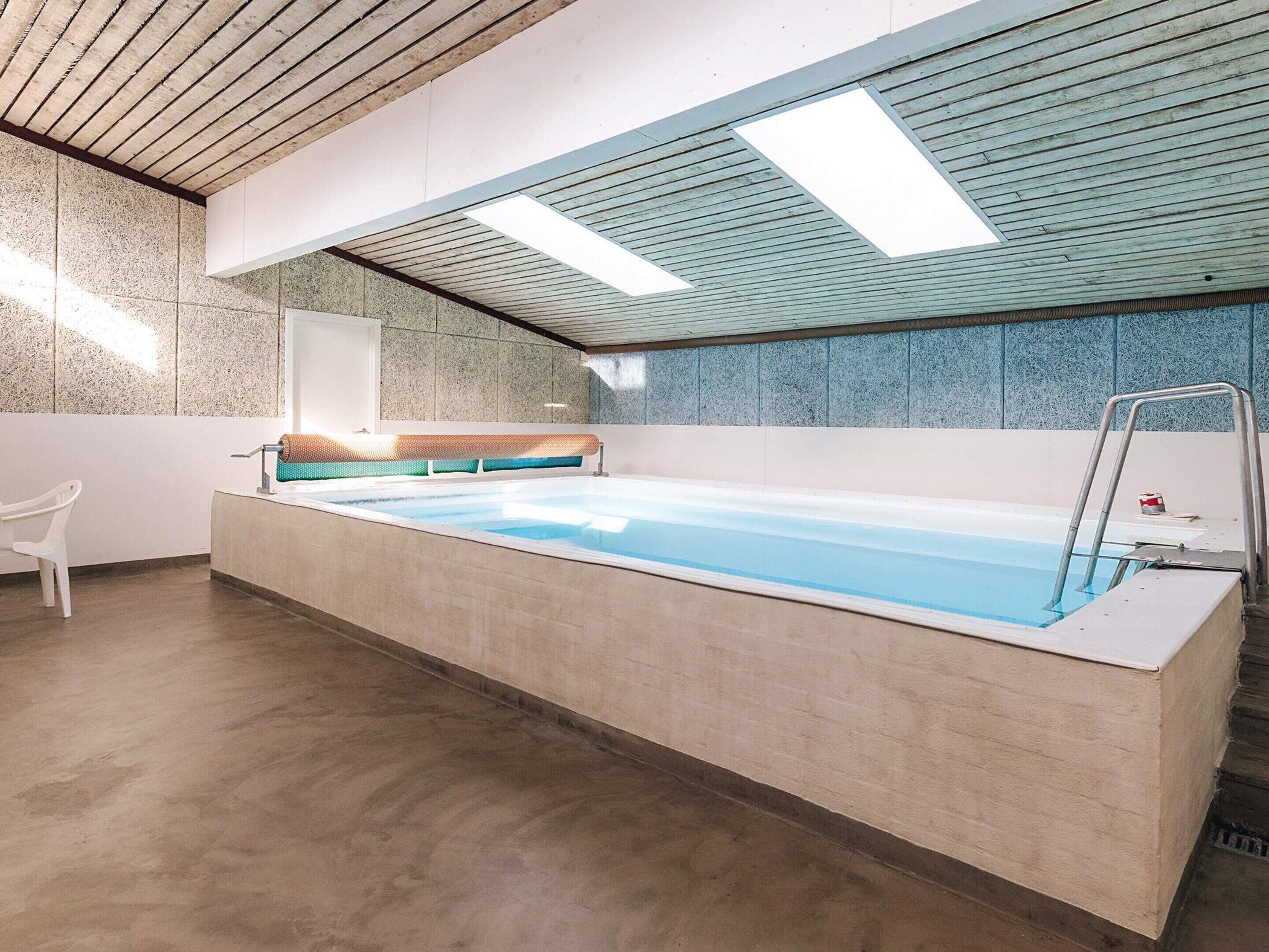 Piscina interior moderna com deck de concreto, teto de madeira e parede revestida em azulejo.