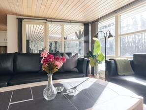 Una sala de estar moderna con un sofá de cuero negro, un florero de vidrio con flores y un ventanal con vistas a árboles.