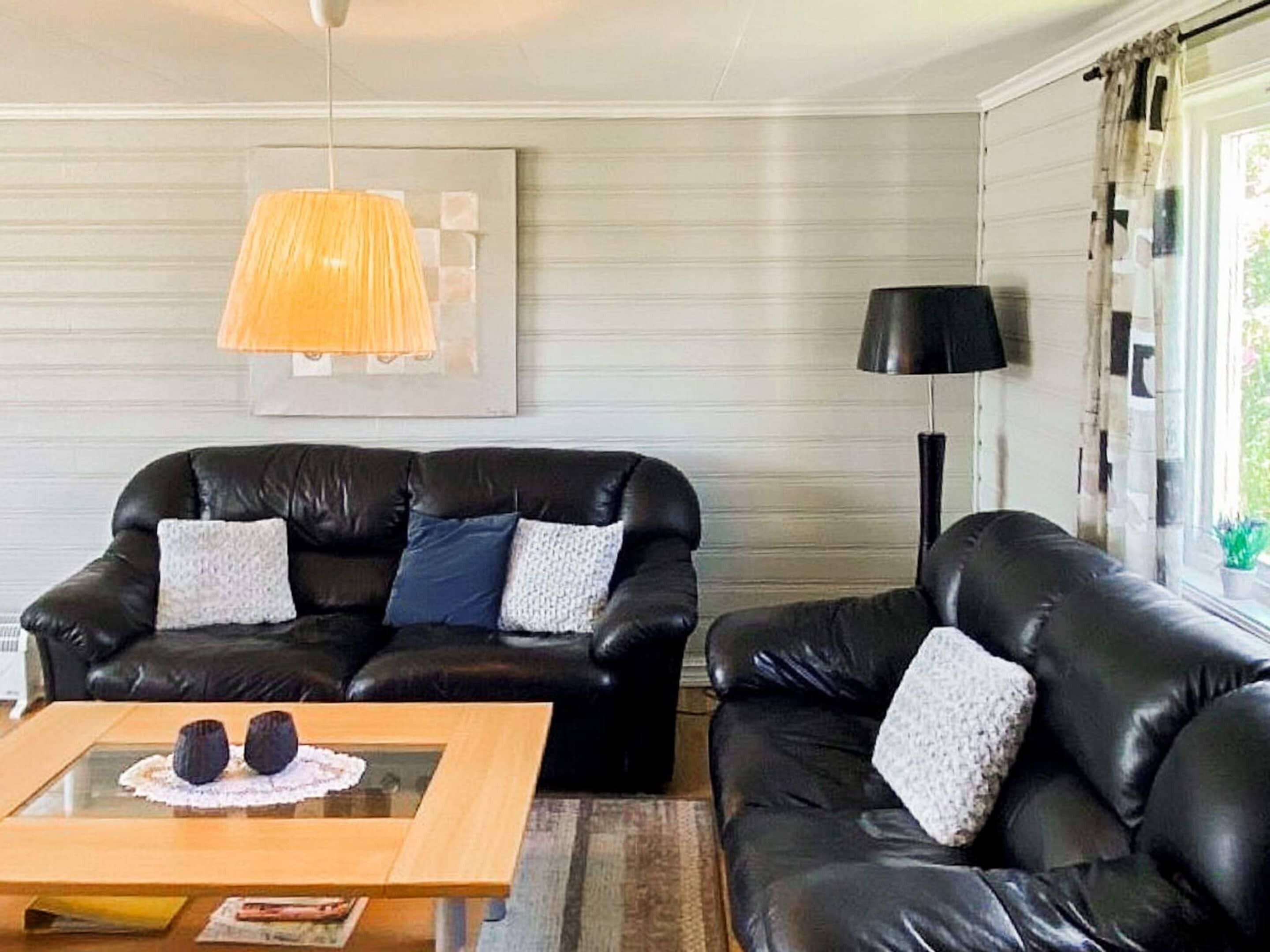 Un salón con un sofá de cuero negro, una mesa de centro de vidrio con objetos decorativos, una lámpara colgante y un cuadro en la pared.