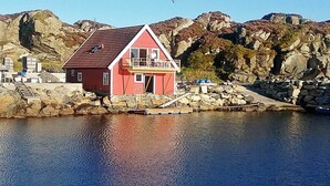 Uma casa vermelha com varanda, localizada junto a uma costa rochosa com um cais que se estende por águas calmas.