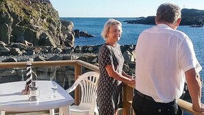 Um casal em frente a uma mesa, com vista para o mar e a costa rochosa.
