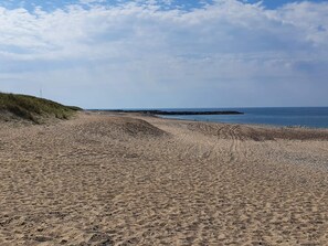 Una playa arenosa con una duna cubierta de hierba, un afloramiento rocoso y un cielo despejado.