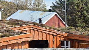 A rustic wooden structure with a stone wall in front, a red barn with a blue roof in the background, and a clear sky. - 4 Star Holiday Home in Fjortoft (Ålesund)