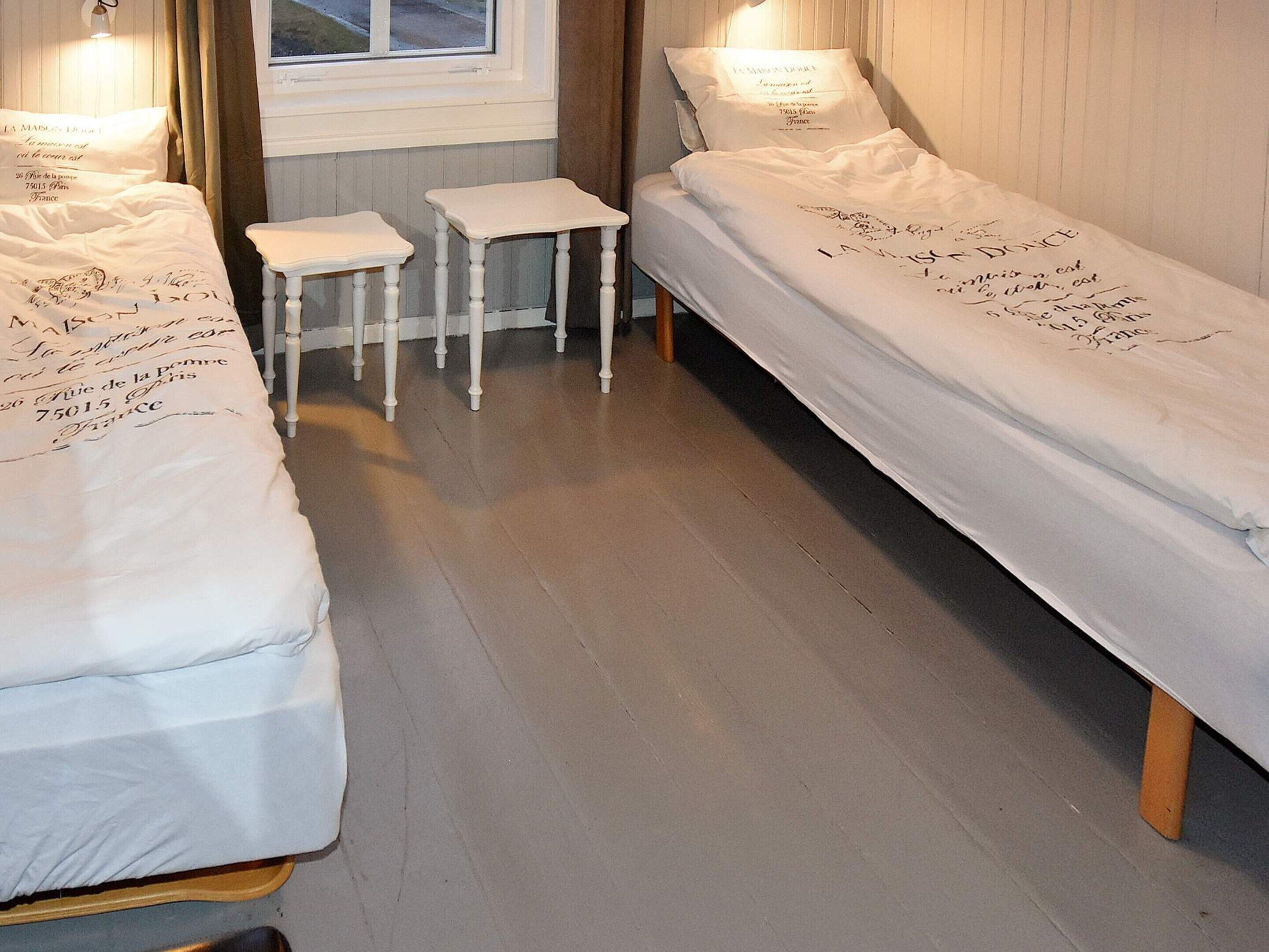 A room with two single beds, each with a white mattress and matching pillows. There are two small white nightstands between the beds.