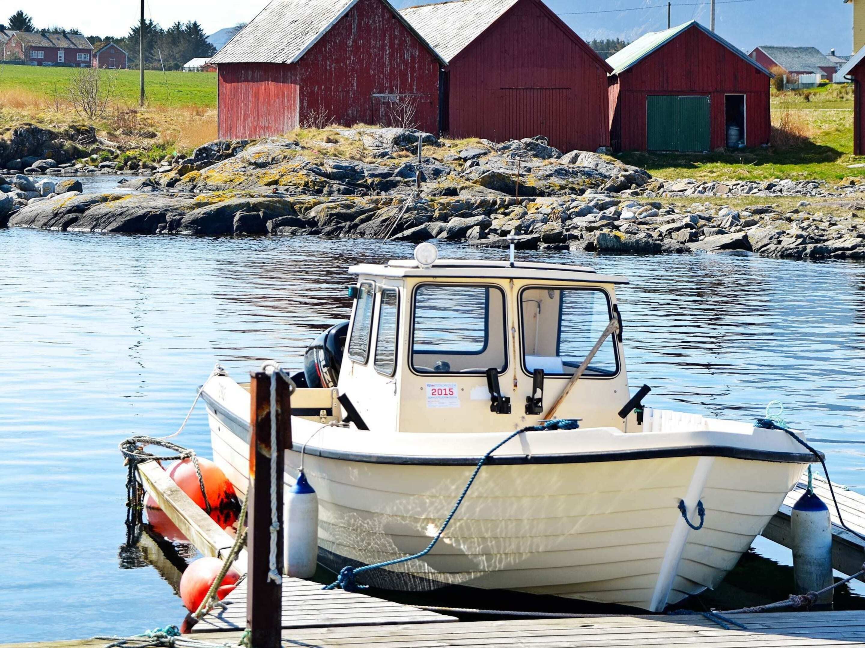 A small boat docked at a wooden pier with a rocky shoreline and red buildings in the background.