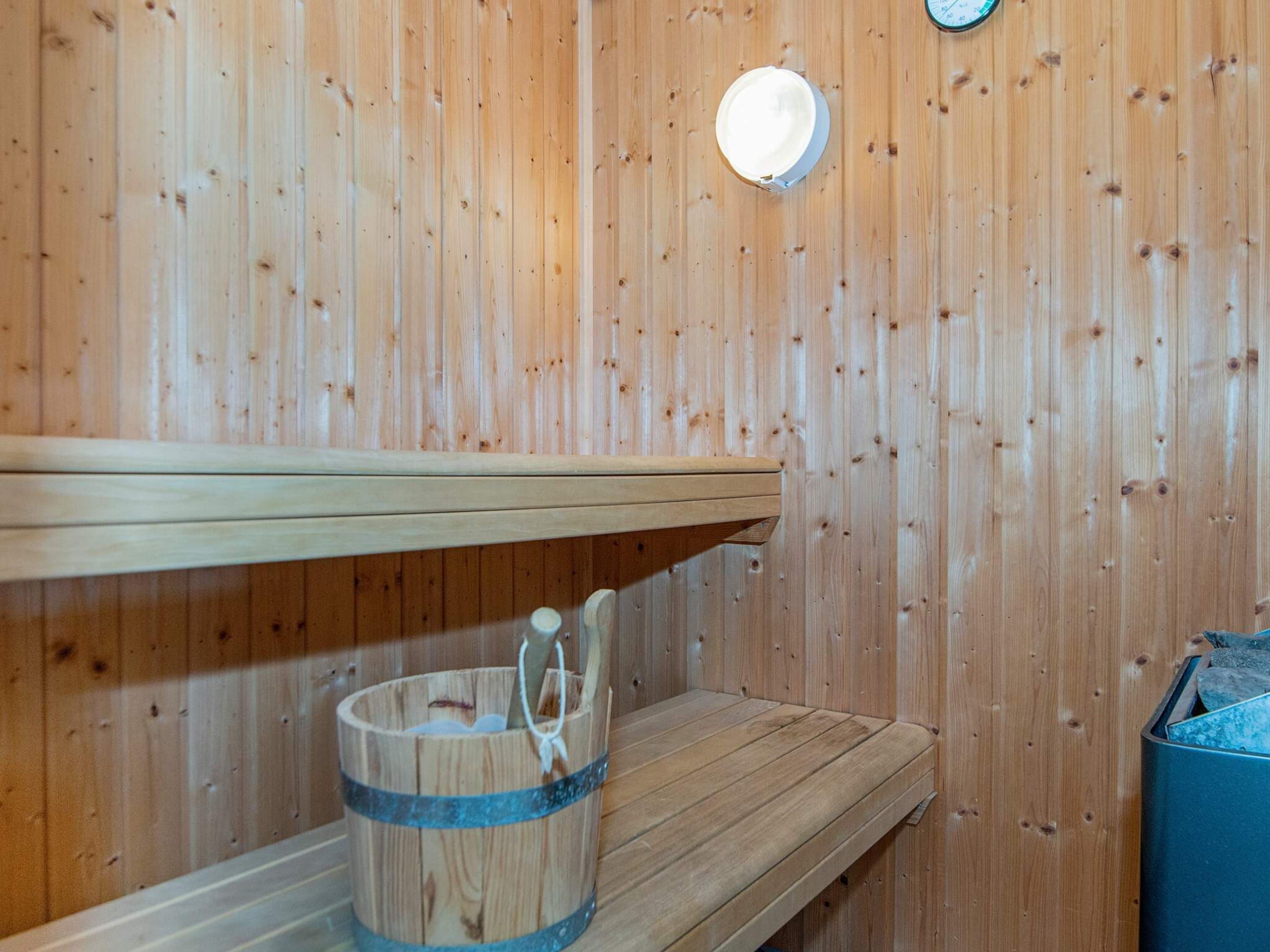 Eine Holzsauna mit einer Sitzbank, einem Eimer mit Bürste und einer Wandleuchte.