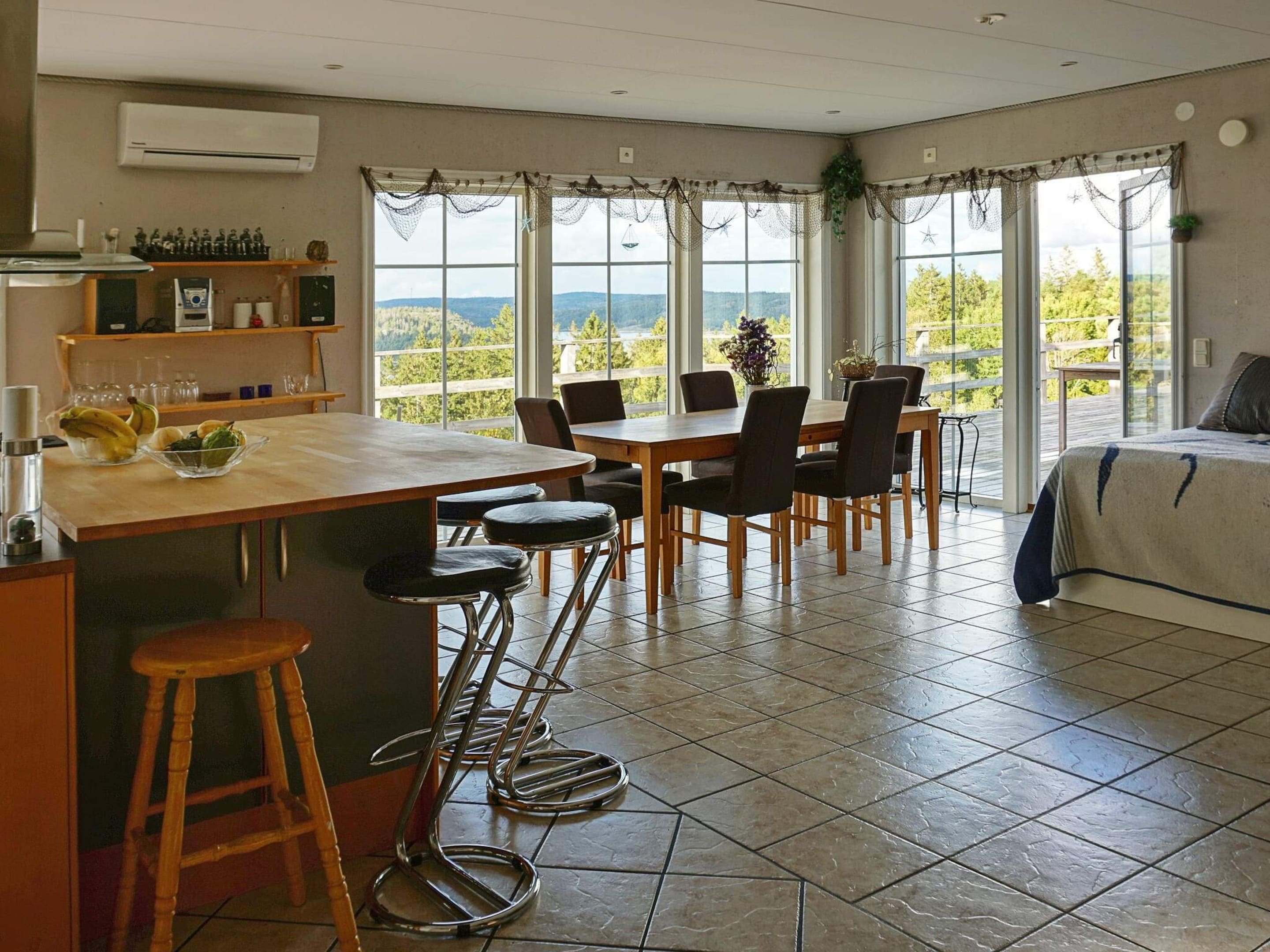 A modern kitchen with a large island, bar stools, and a dining area with chairs. There are large windows with a view of the outdoors.