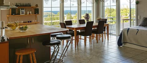 A modern kitchen with a large island, bar stools, and a dining area with chairs. There are large windows with a view of the outdoors.