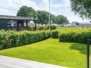 Une pelouse bien entretenue, agrémentée d’une terrasse en bois, le tout entouré de haies et d’arbres taillés avec soin.