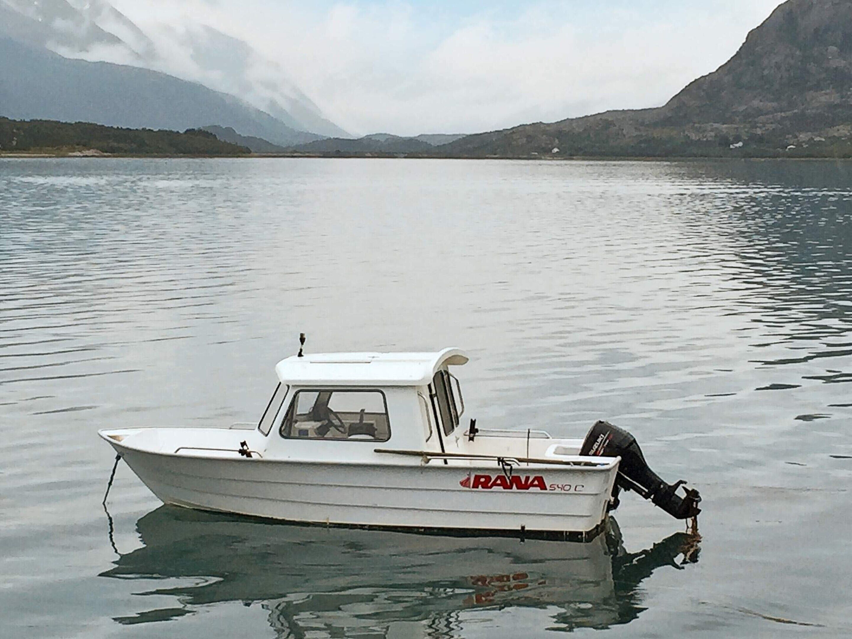Ein weißes Boot namens "RANA" auf ruhigem Wasser, mit Bergen im Hintergrund.