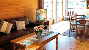 Een gezellige woonkamer met een houten vloer, een bank, een salontafel met een bloevas, een televisie en een eethoek met stoelen.