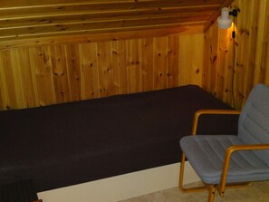 A single bed with a dark mattress in a room with wooden walls and a single chair with a blue cushion. - 6 Person Holiday Home in Torsby (Torsby)