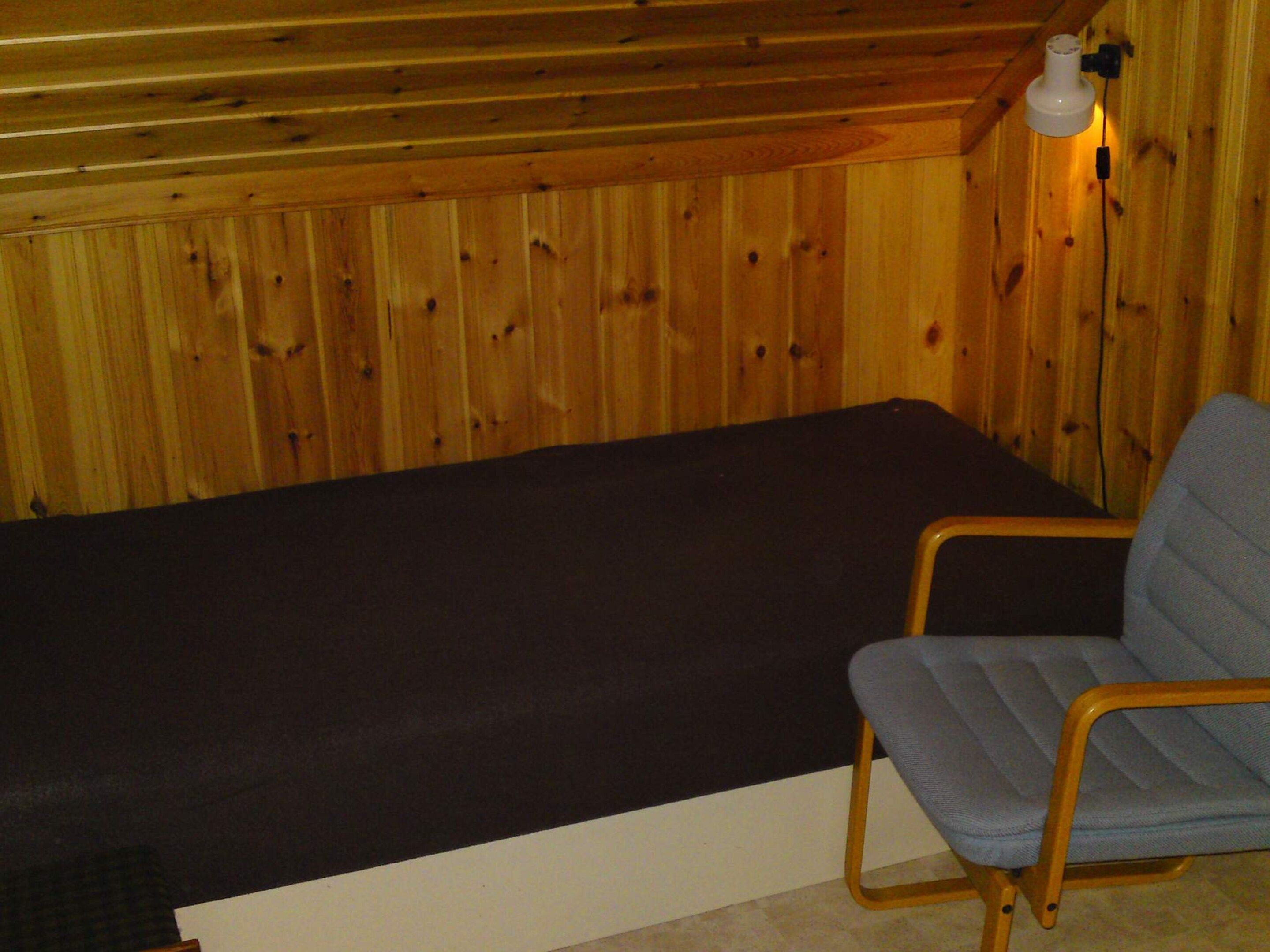 Uma cama de solteiro com colchão escuro em quarto com paredes de madeira e uma única cadeira.
