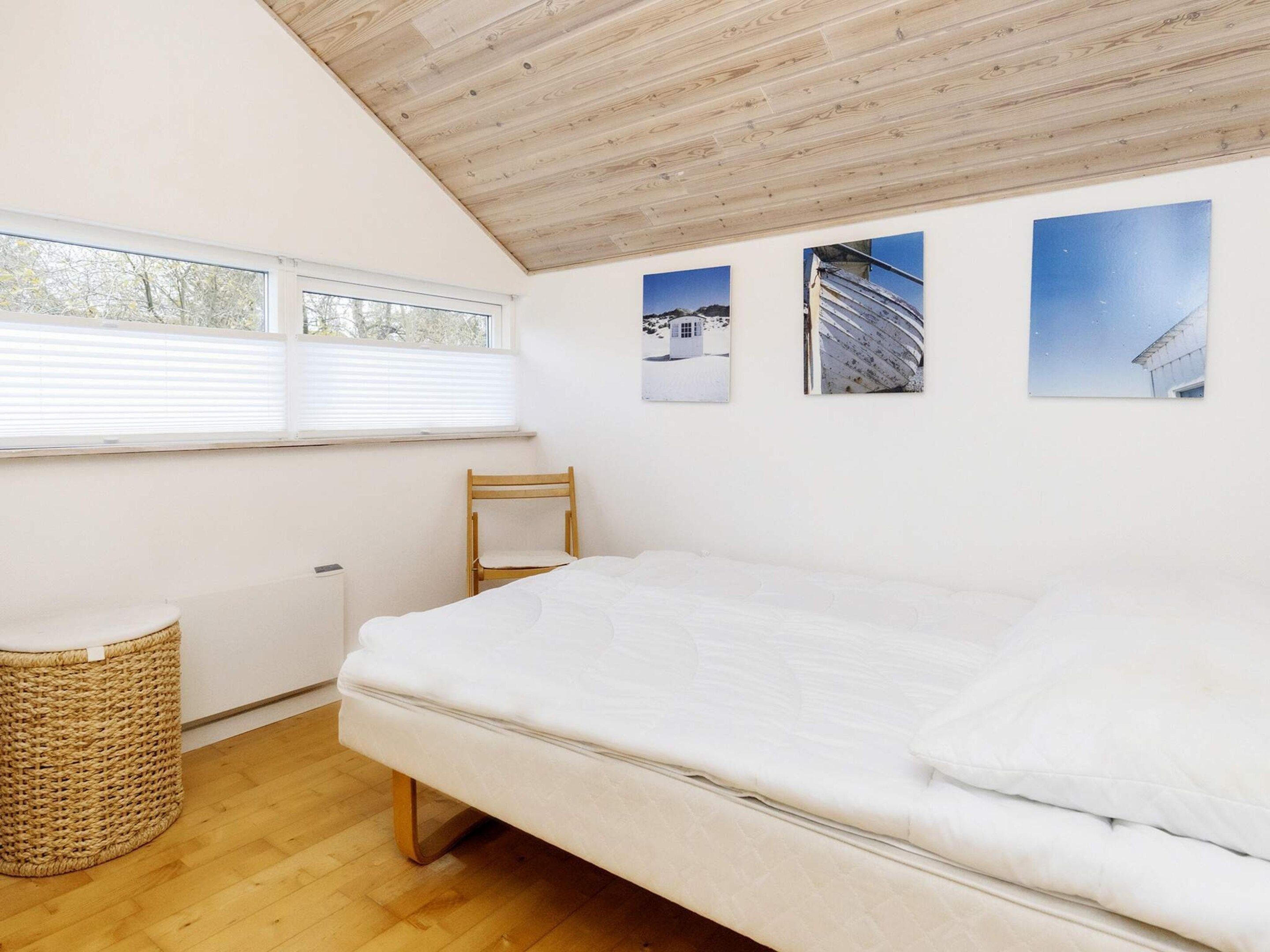 A bedroom with a wooden ceiling, a bed, a chair, a window, and three framed pictures on the wall.