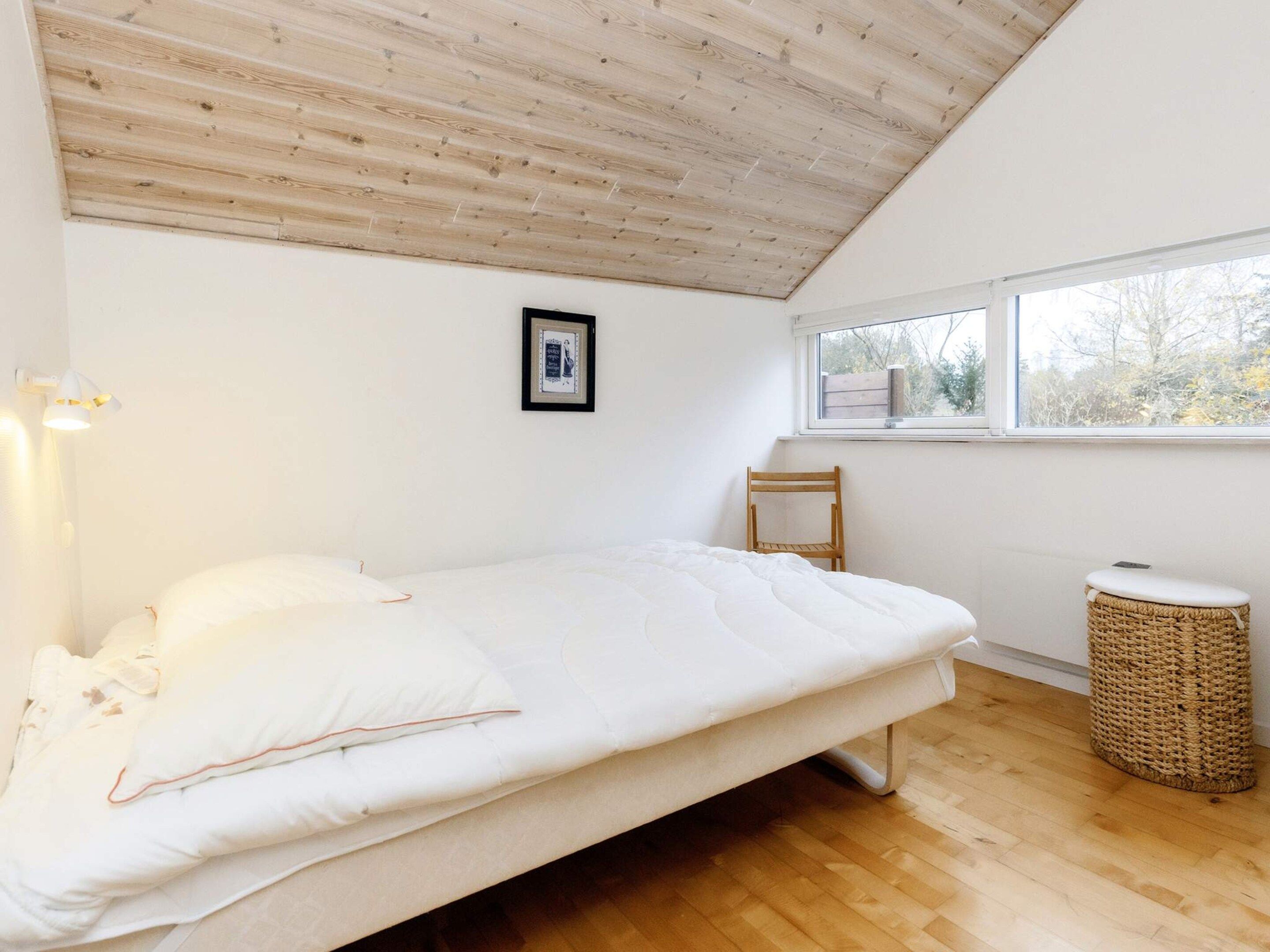 Une chambre avec un plafond en bois, un lit avec une literie blanche, une petite chaise, une fenêtre et un panier en osier.