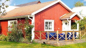 Une maison en bois rouge avec une fenêtre blanche, une terrasse en bois avec des bancs bleus et un jardin où poussent différentes plantes.