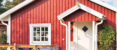 Un bâtiment en bois rouge avec une porte et une fenêtre blanches, un porche avec des bancs bleus et un petit jardin.