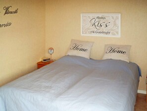 Ein Schlafzimmer mit einem großen Bett, zwei Kissen mit dem Aufdruck 'Home', einem Nachttisch mit Lampe und Radio sowie einer Wanddekoration mit der Aufschrift 'Always Kiss me Goodnight'.