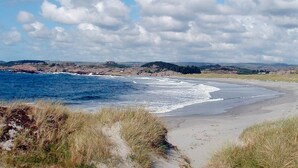 Paisaje costero con una playa de arena, dunas cubiertas de hierba y vista al océano bajo un cielo parcialmente nublado.