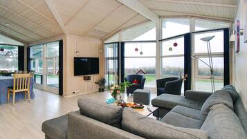 A spacious living room with a large grey sectional sofa, a television mounted on the wall, and a dining area with a table and chairs.