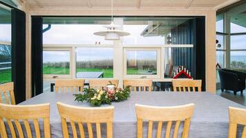 A dining area with a large table and wooden chairs, large windows with a view of the outdoors, and a hanging light fixture.