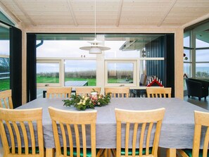 A dining area with a large table and wooden chairs, large windows with a view of the outdoors, and a hanging light fixture.