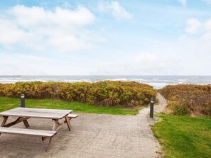 A picnic area with a wooden table and benches, a paved path leading to the sea, and a hedge in the foreground. - 5 Star Holiday Home in Gorlev (Gørlev)