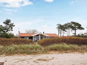 Une maison situĂ©e sur la cĂŽte, avec un toit rouge, de grandes fenĂȘtres et une plage de sable au premier plan.