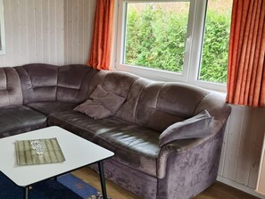 Um sofá em pele castanha, tipo sectional, com uma mesa de centro branca à frente, posicionado ao lado de uma janela com cortinas laranja.