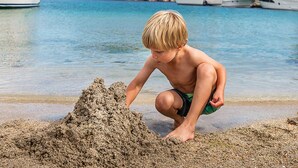 Et barn leger i sanden på en strand med både og klipper i baggrunden.