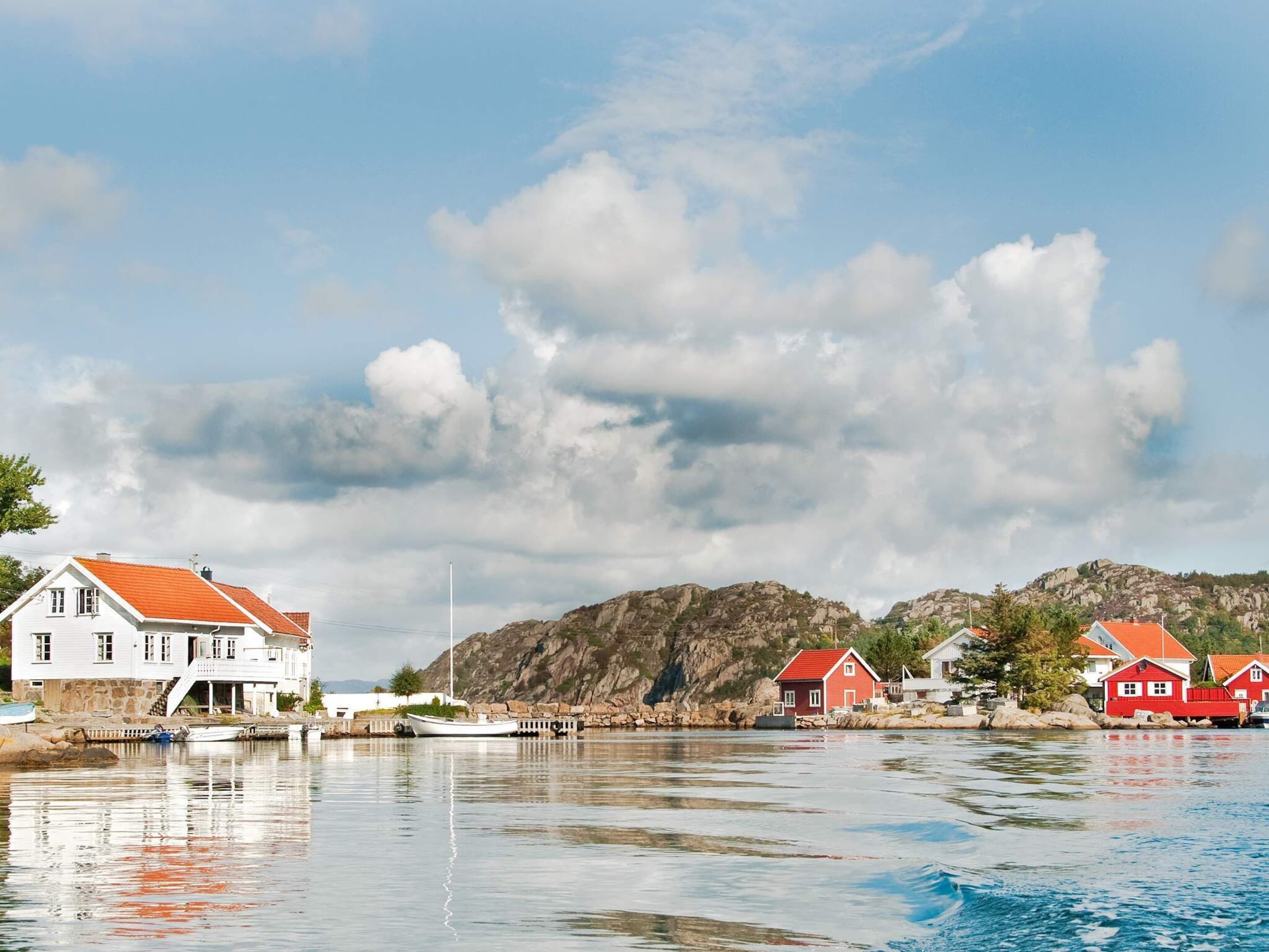 A coastal scene with a white building with a red roof, a red house, and rocky hills in the background.