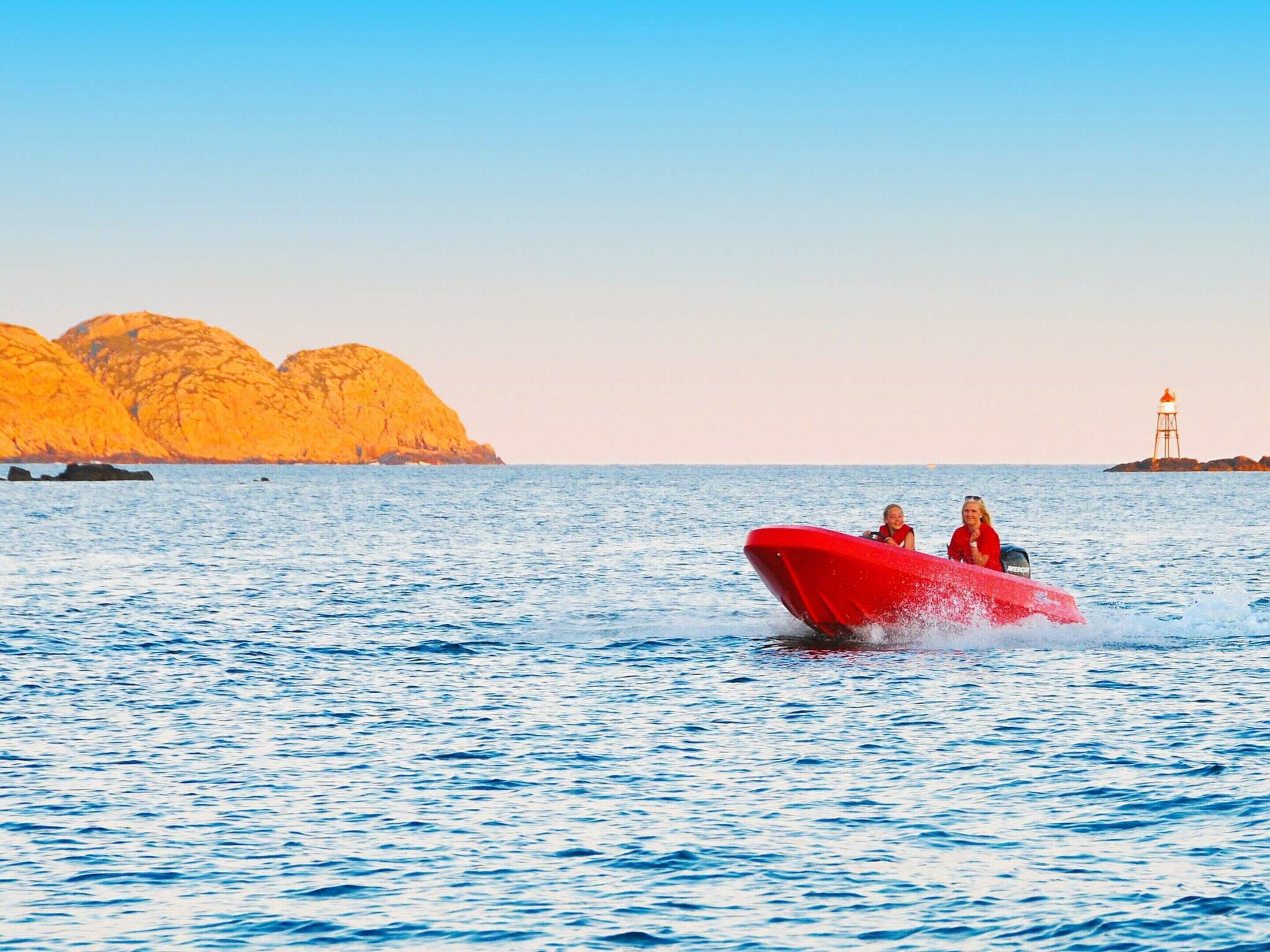 A red speedboat on calm water with rocky cliffs and a lighthouse in the distance.