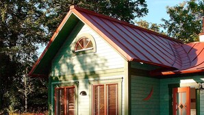 A small, single-story house with a green exterior and a red roof. It features a front porch with a wooden table and chairs.