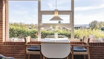 Comedor con mesa y sillas, lámpara colgante y vistas a zonas verdes y casas a través de amplios ventanales.