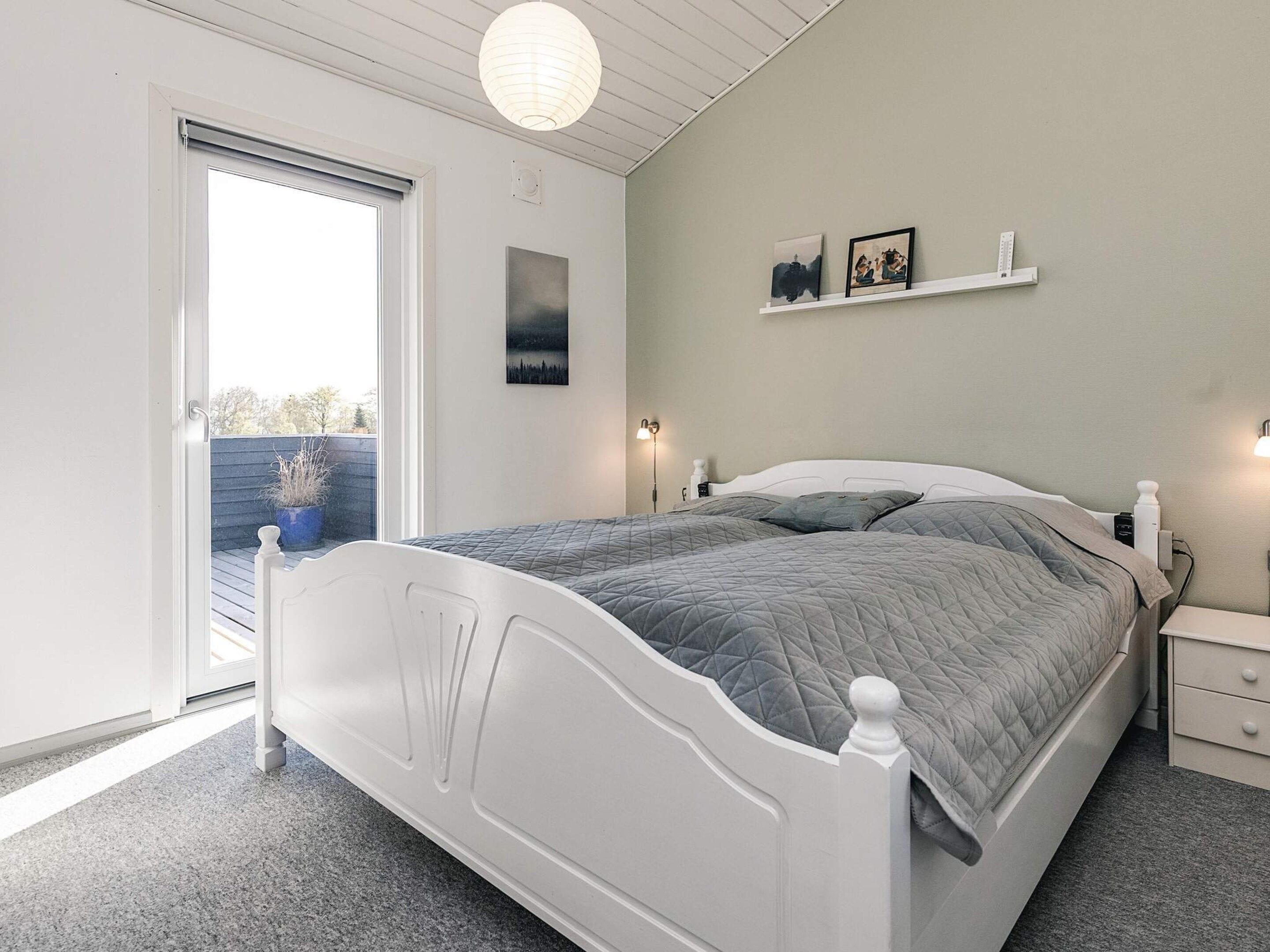 Un dormitorio con una cama blanca, una mesita de noche, una repisa en la pared con fotografías y una ventana con vistas al exterior.