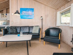 Et hyggeligt rum med en sort lædersofa, et hvidt bord og en sort læderstol. Et maleri af et slot hænger på væggen, og der er et vindue med gardiner.