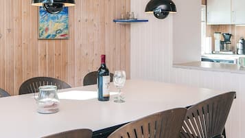 A dining area with a table set for two, a bottle of wine, and two glasses. There are two black pendant lights above the table, and a wooden wall with a colorful painting. A kitchen area is visible in the background.