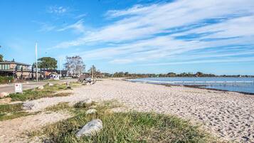 A sandy beach with a clear sky, a few buildings in the background, and a calm body of water.