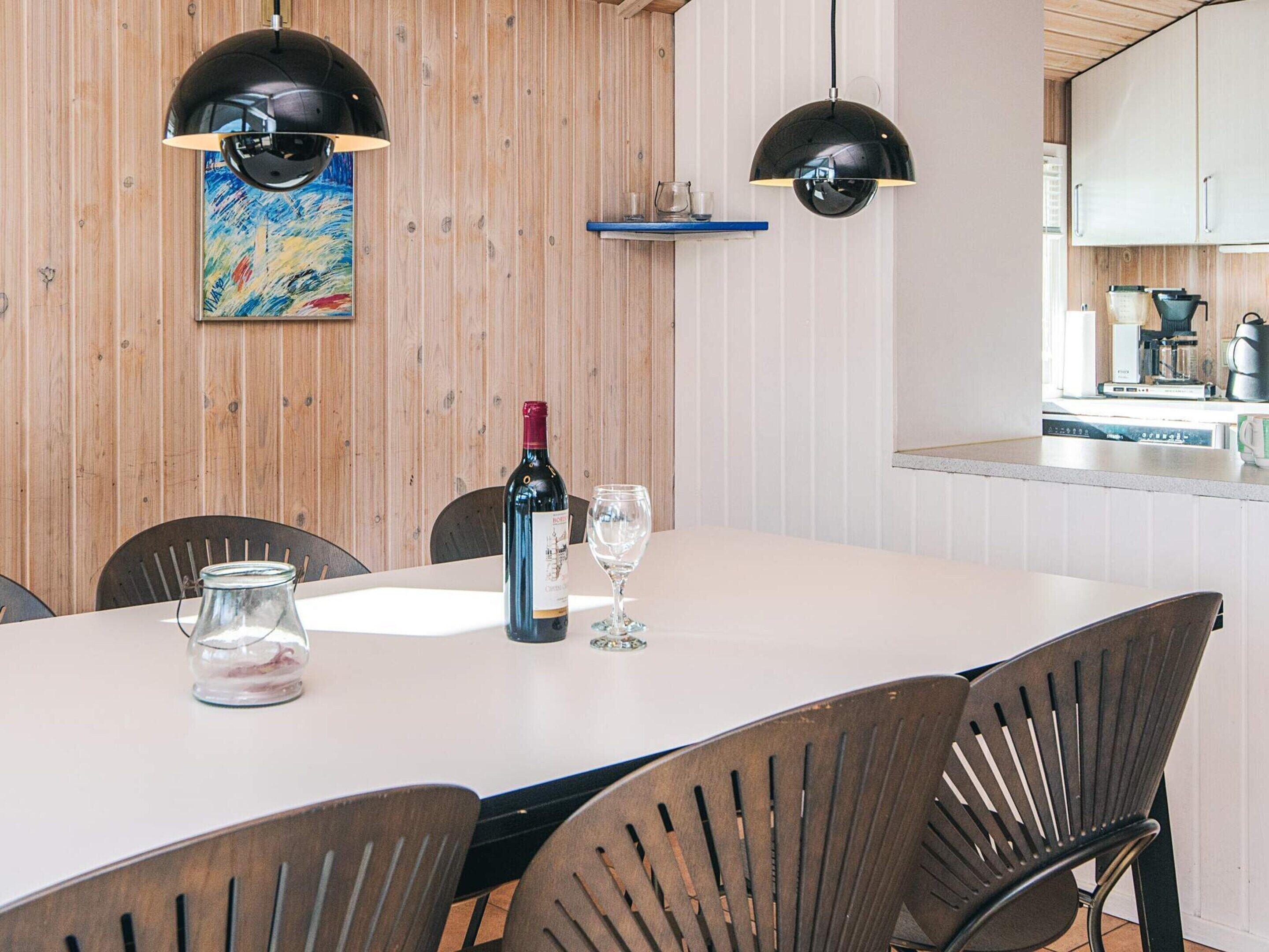 A dining area with a table set for two, a bottle of wine, and two glasses. There are two black pendant lights above the table, and a wooden wall with a colorful painting. A kitchen area is visible in the background.