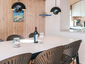 A dining area with a table set for two, a bottle of wine, and two glasses. There are two black pendant lights above the table, and a wooden wall with a colorful painting. A kitchen area is visible in the background. - 4 Star Holiday Home in Bjert (Bjert)