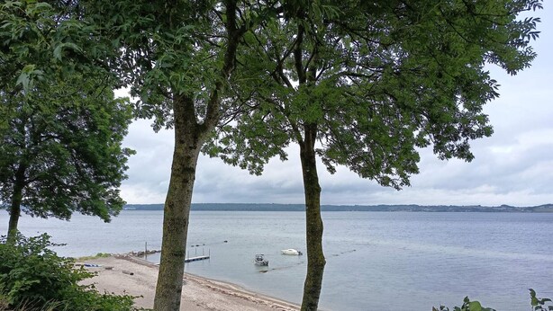 A lakeside view with trees, a sandy beach, and a small boat. - 6 Person Holiday Home in Aabenraa (Aabenraa)