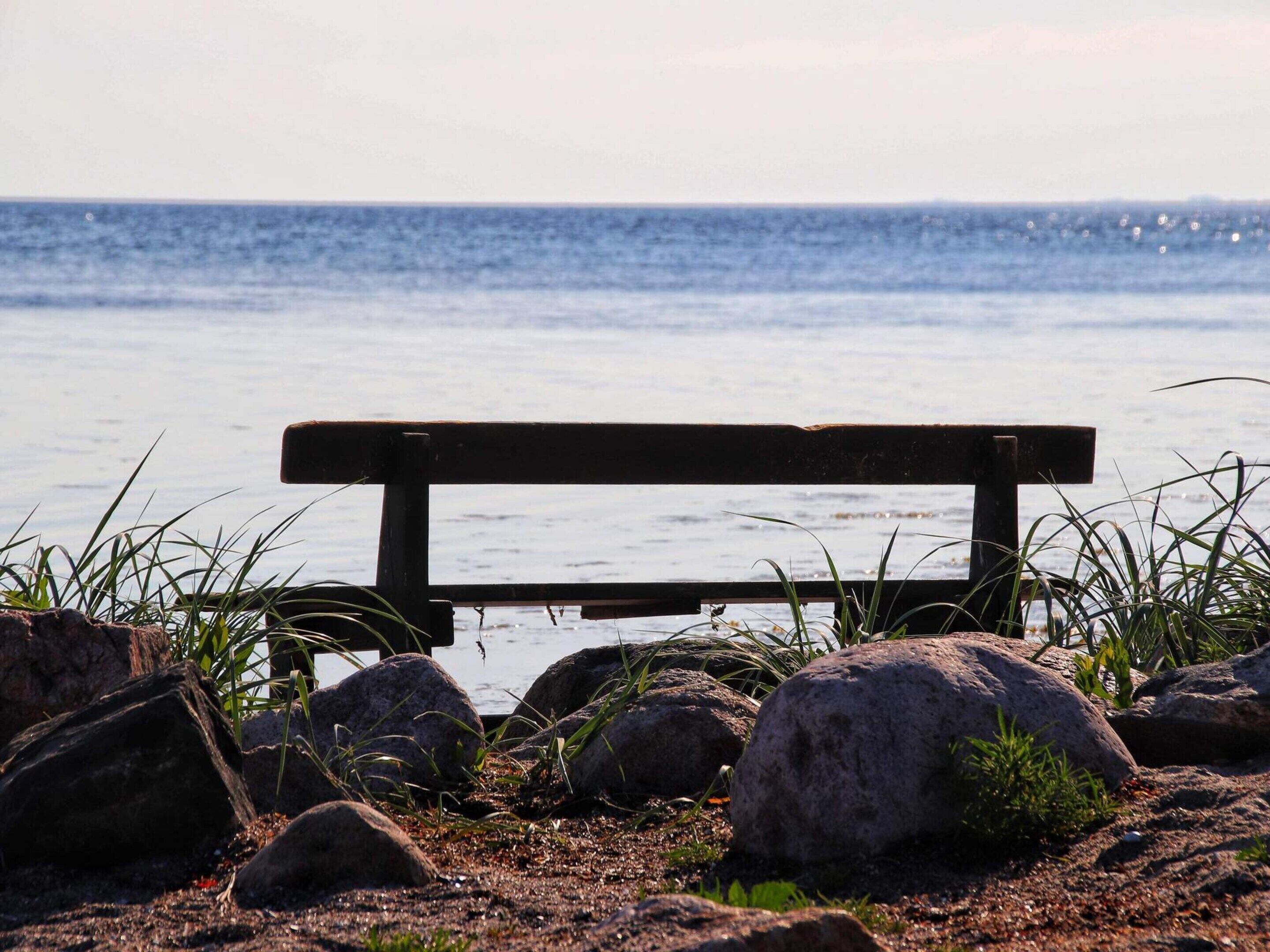 Un banc au bord de l’eau, avec des rochers et de l’herbe au premier plan.