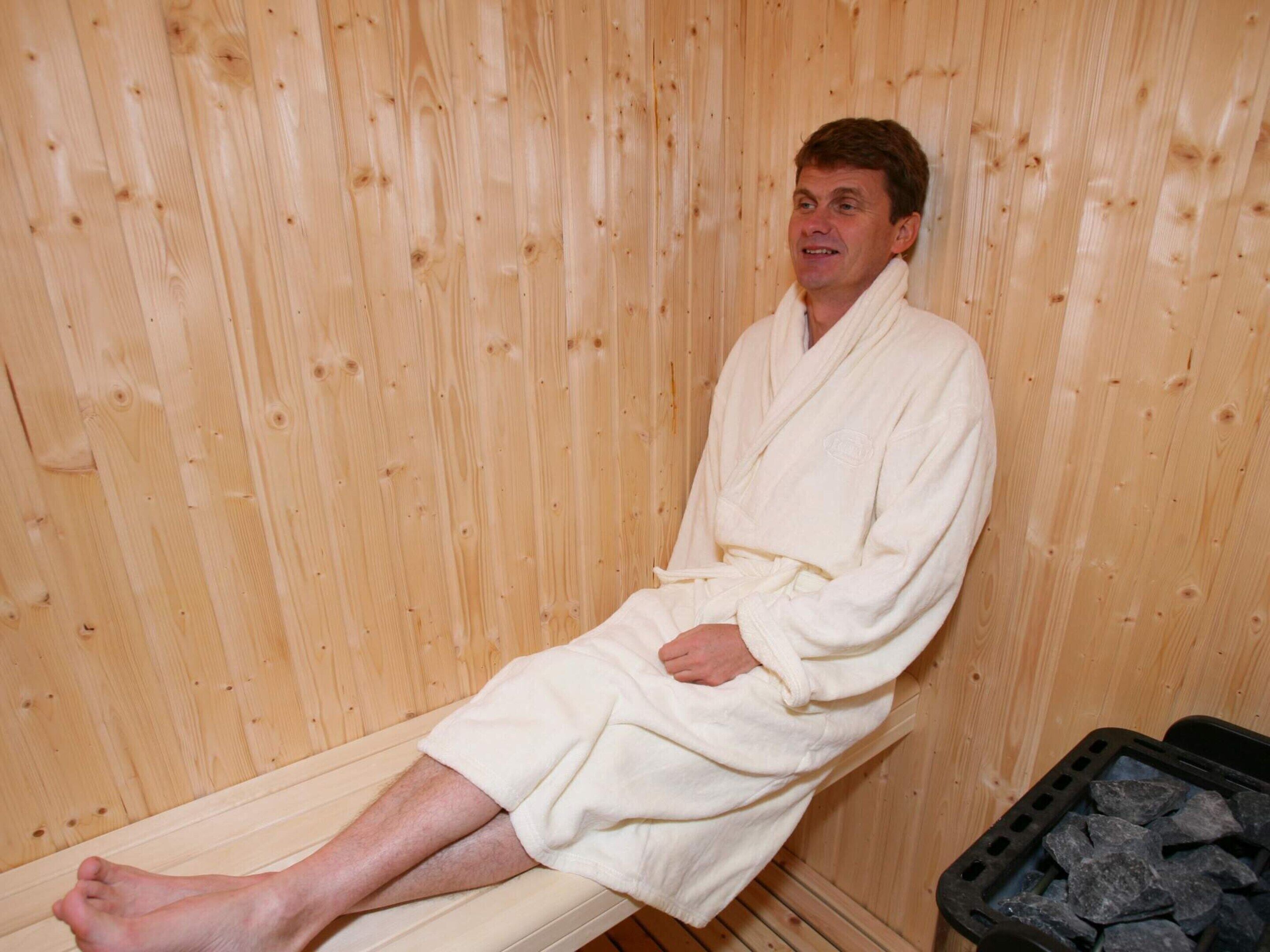 A person seated in a wooden sauna with a bench and a container of stones.