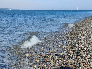 Ein kiesiger Strand mit klarem Wasser und entfernten Segelbooten.