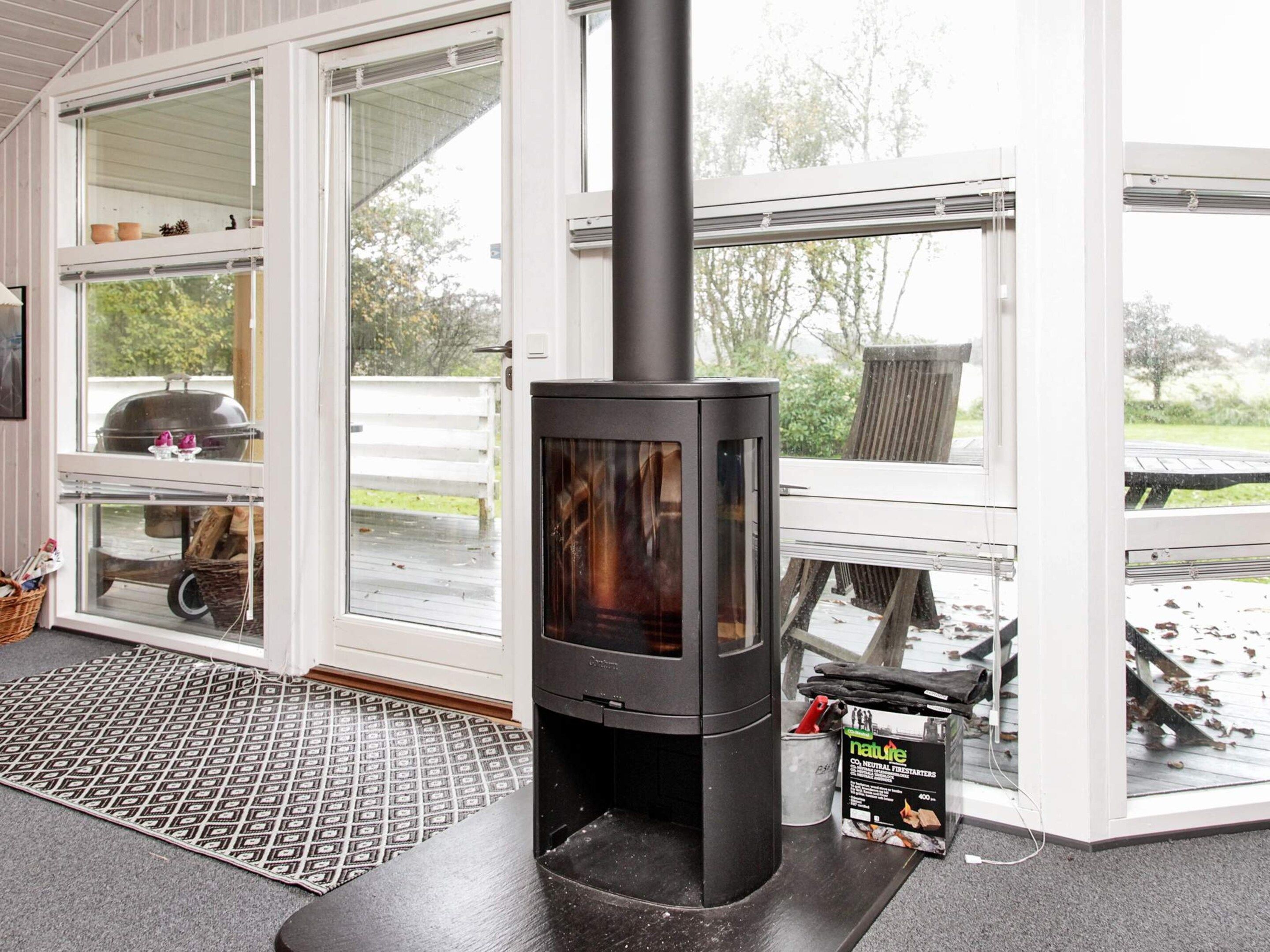 A wood-burning stove in a room with large windows, a grill outside, and a mat on the floor.